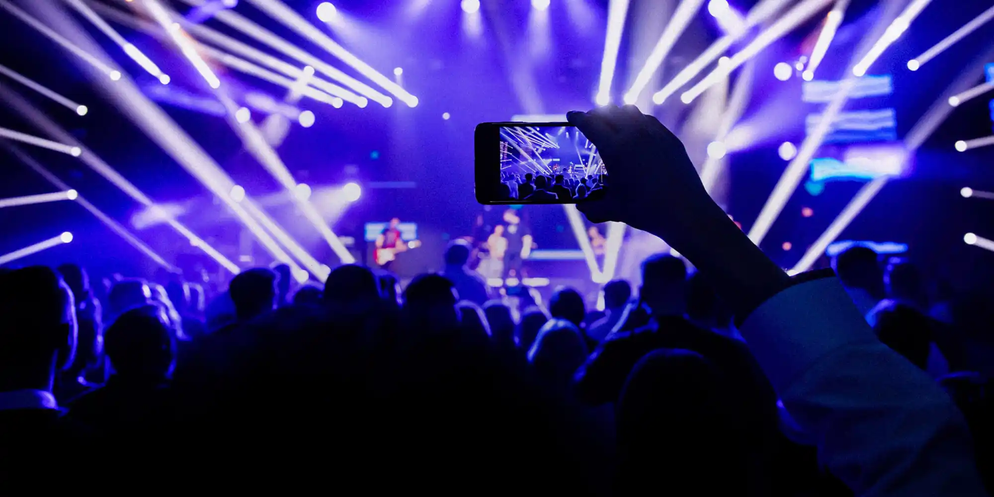 An outstretched hand over a crowded audience holding a smartphone and recording video of a concert