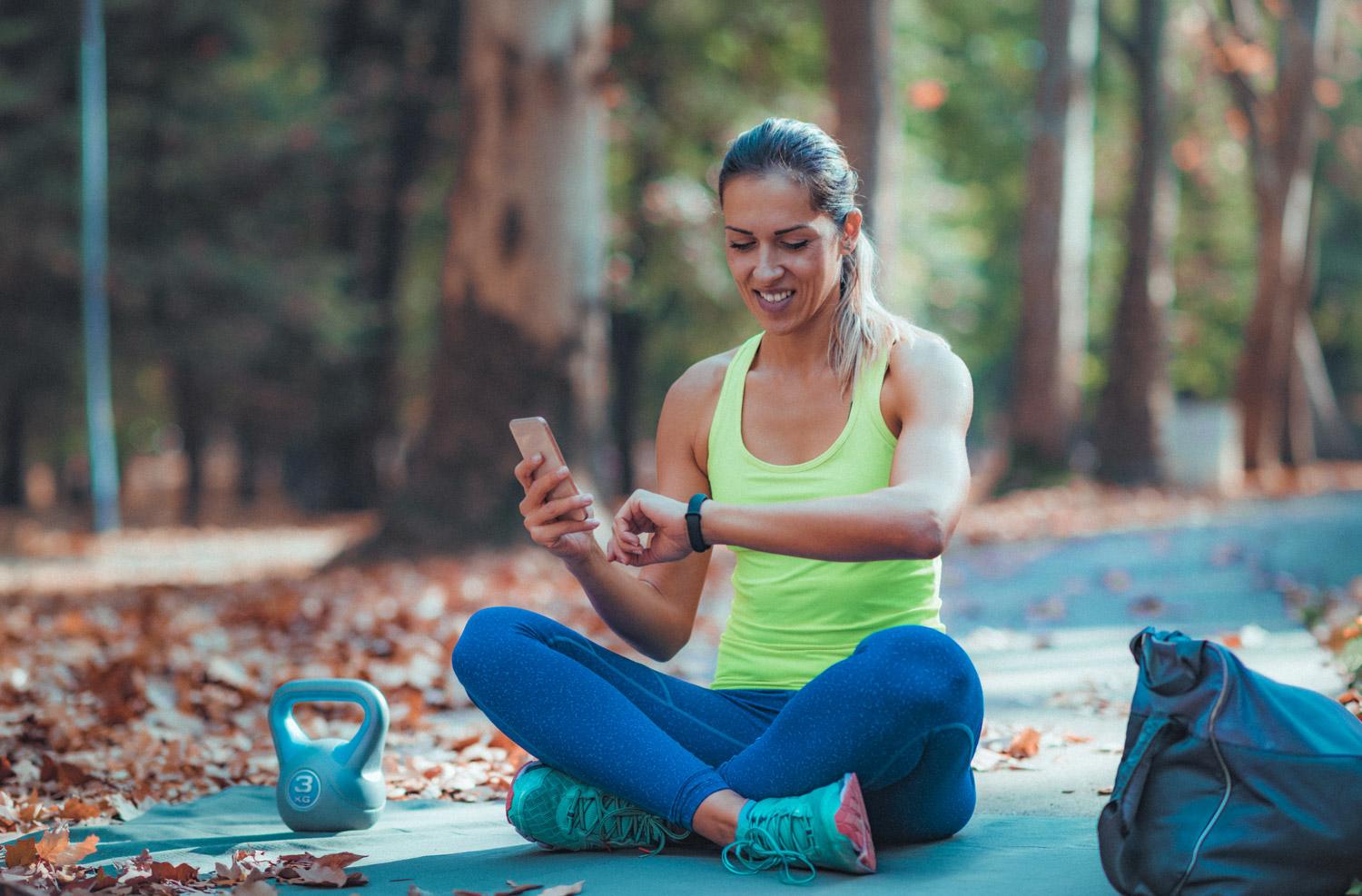 Photograph of a woman sitting on a yoga mat in a forest looking at the wearable tech device on her wrist