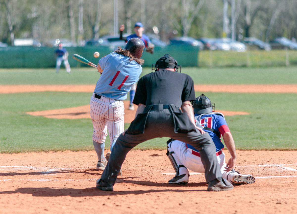 Photograph of a batter hitting a ball in a baseball game, from behind the umpire’s perspective