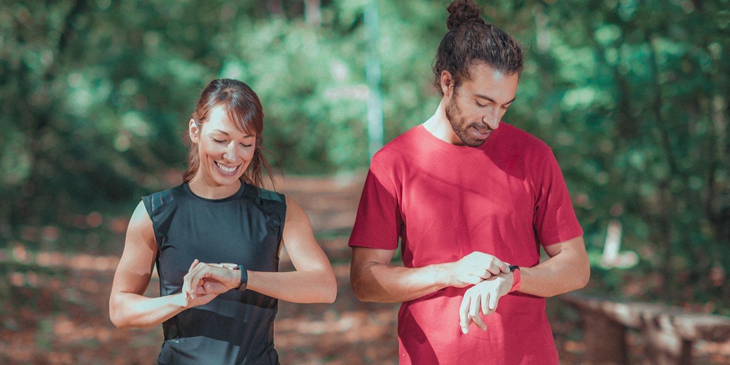 Photograph of a woman and a man looking at their wrist watches, with a forest trail in the background