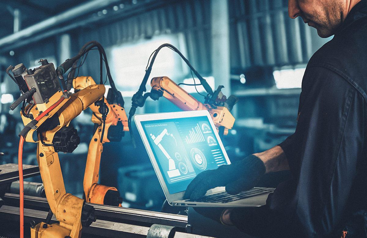 Photograph of man in a manufacturing facility working on a laptop with robotic arms behind it