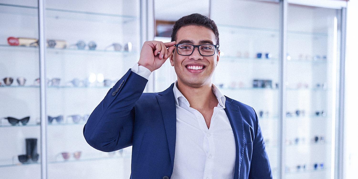 Photo of a person adjusting his glasses in front of a wall of different types of eyeglasses