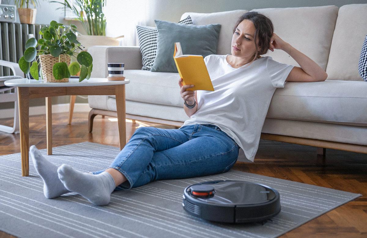 Photograph of woman reading a book while sitting on the floor of a living room, as a robotic vacuum cleaner works beside her
