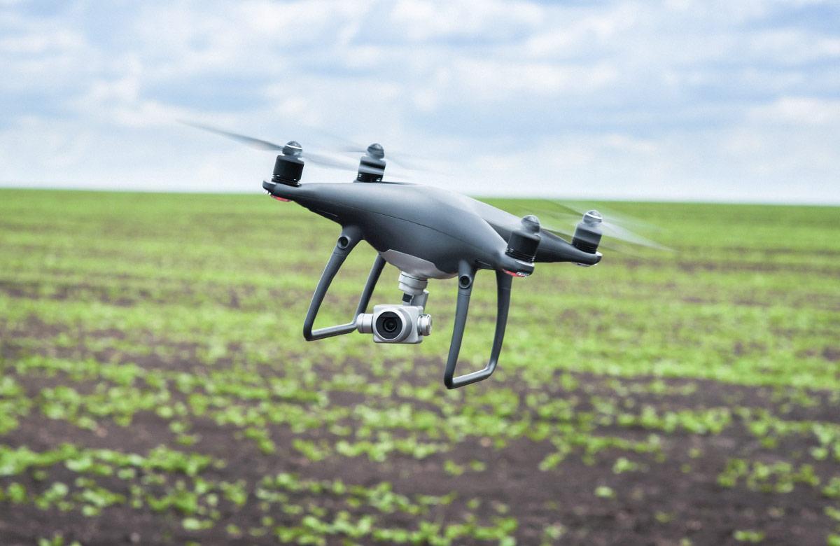 Gray drone in flight over a green field of crops under a cloudy sky, illustrating agricultural technology.