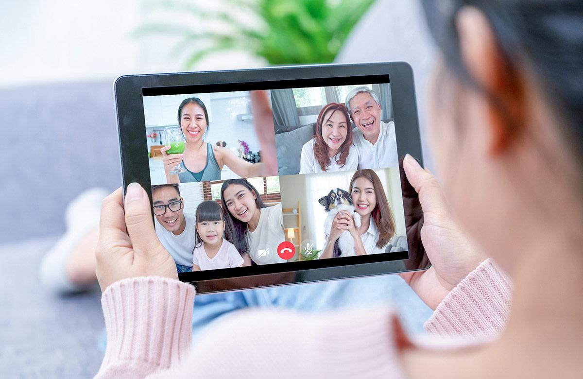 Woman using a tablet for a video call with multiple family members, including smiling elderly couple, parents with a child, and a woman holding a dog.