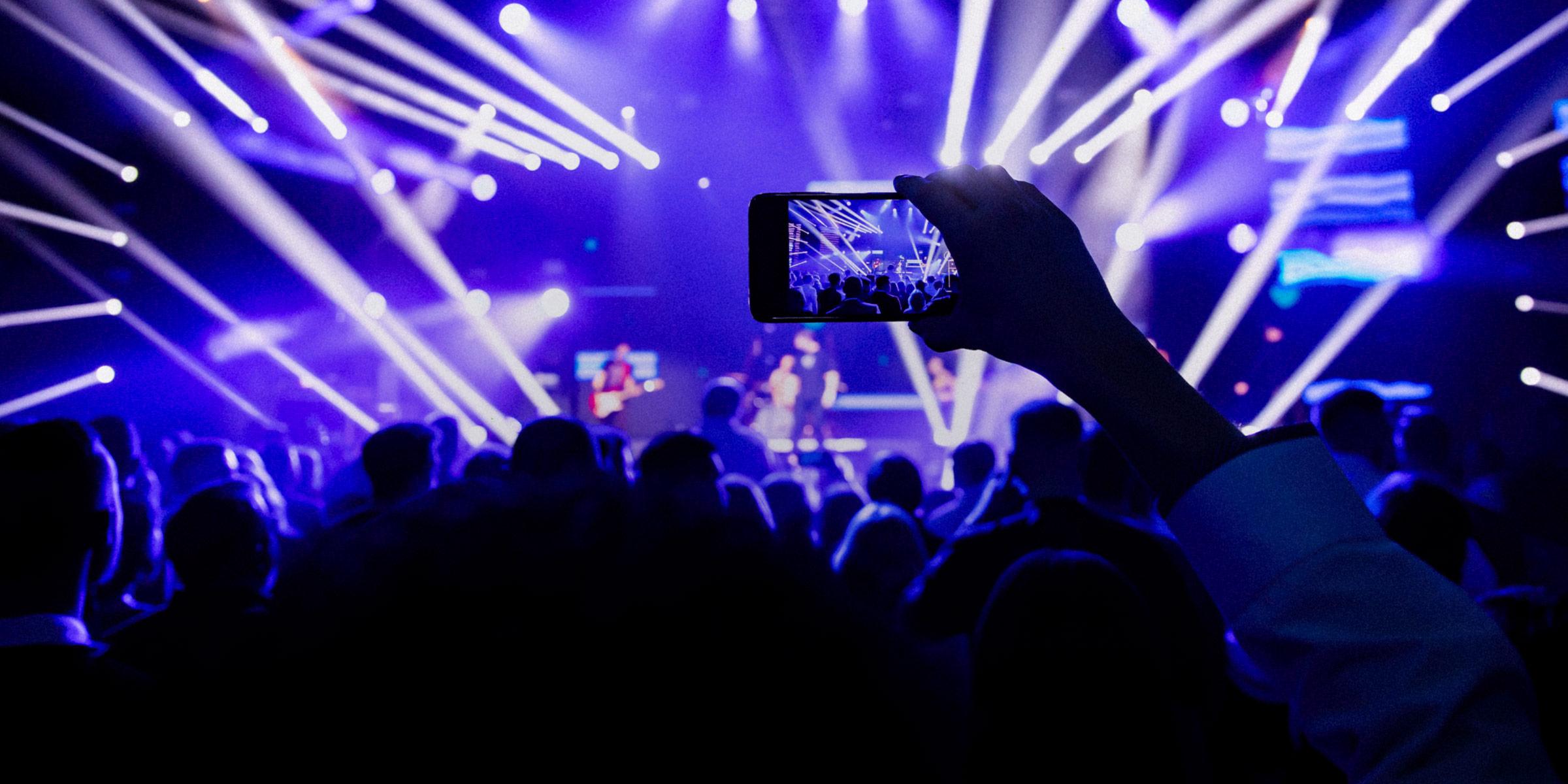 An outstretched hand over a crowded audience holding a smartphone and recording video of a concert.