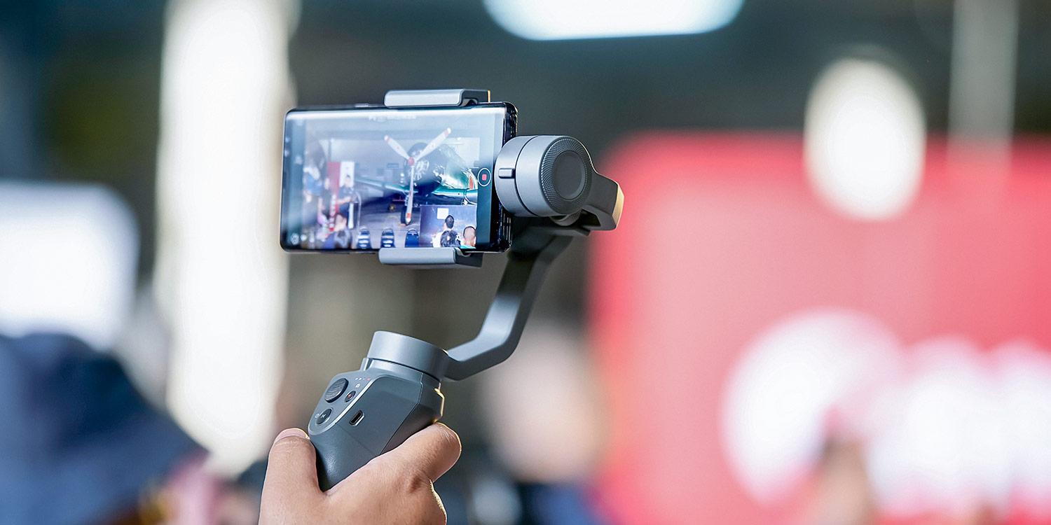 A close-up shot of a smartphone mounted on a gray handheld gimbal, held by a person's hand with blurred background elements. The smartphone screen displays a video, likely a live feed, with various people and a small airplane visible. The gimbal features buttons and controls on the handle. The background is out of focus, featuring light and a red backdrop.