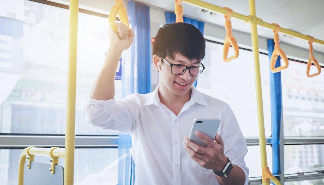 Young man wearing glasses and ear bud headphones looks down at smartphone while standing on public transportation holding hand rail