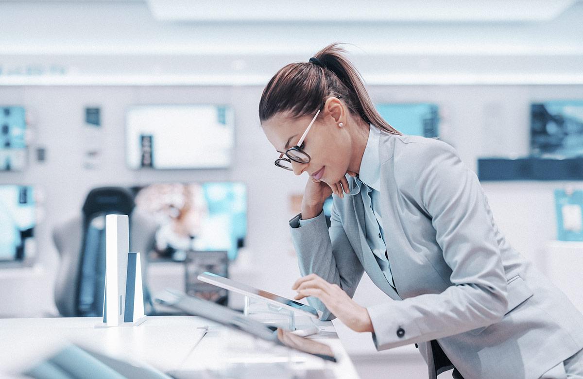 A businesswoman in a light gray suit and glasses leans over a tablet in a tech store, looking at the screen with one hand on her face. Other tablets are on the display, and TV screens are visible in the background.