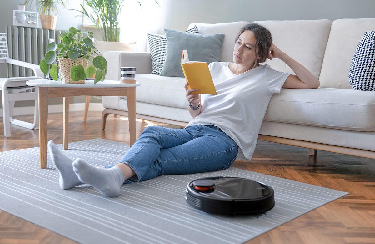 Woman sitting on floor reading a book while a robotic vacuum cleaner works in the foreground