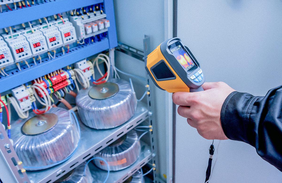Person using a handheld thermal imaging camera to inspect the temperature of electrical components inside an open control panel.