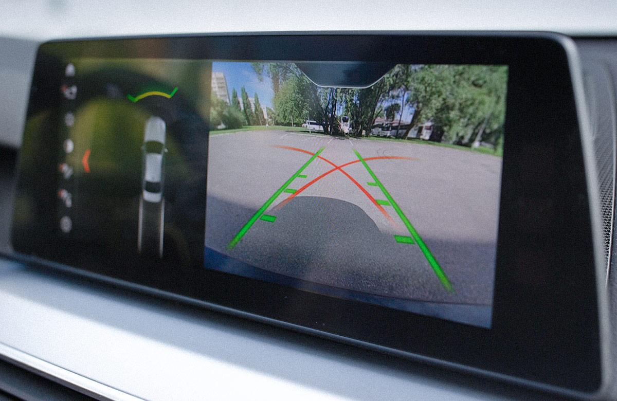 Car dashboard display showing a rearview camera image with colored guide lines, indicating the area behind the vehicle for parking assistance.