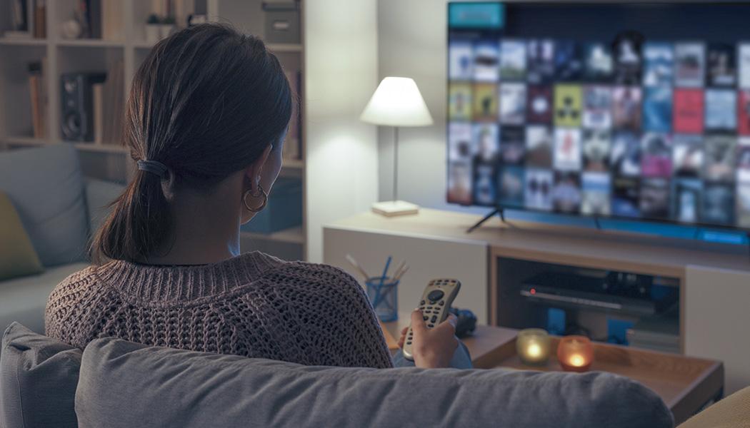 Over-the-shoulder view of woman holding smart TV remote while watching tv in dimly lit room