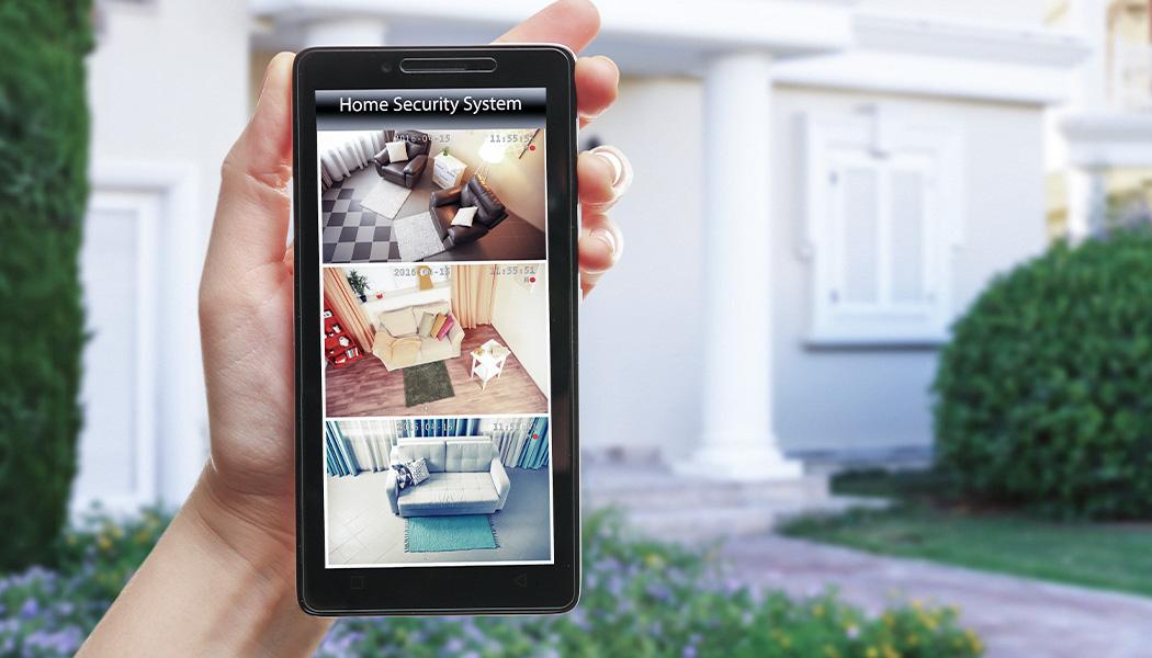 Close-up of hand holding a smartphone on blurred house background. Smartphone display shows video preview of a home security system in a three panel view