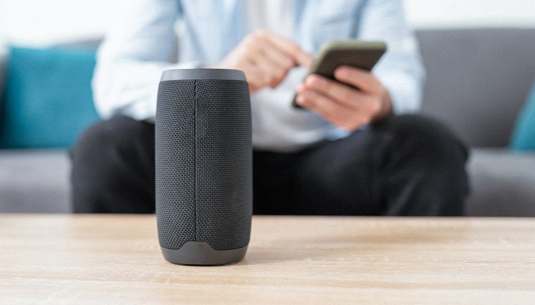 Man in background out of focus, sitting on couch connecting a phone and smart speaker which is shown in foreground of image in focus